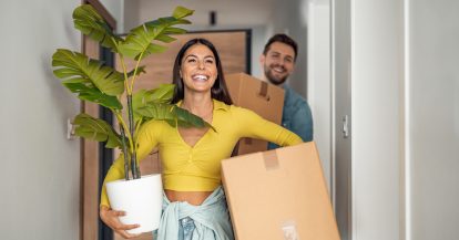 Smiling woman in yellow shirt holding large potted plant in one hand and a box in the other with a man in a blue shirt carrying a box in the background