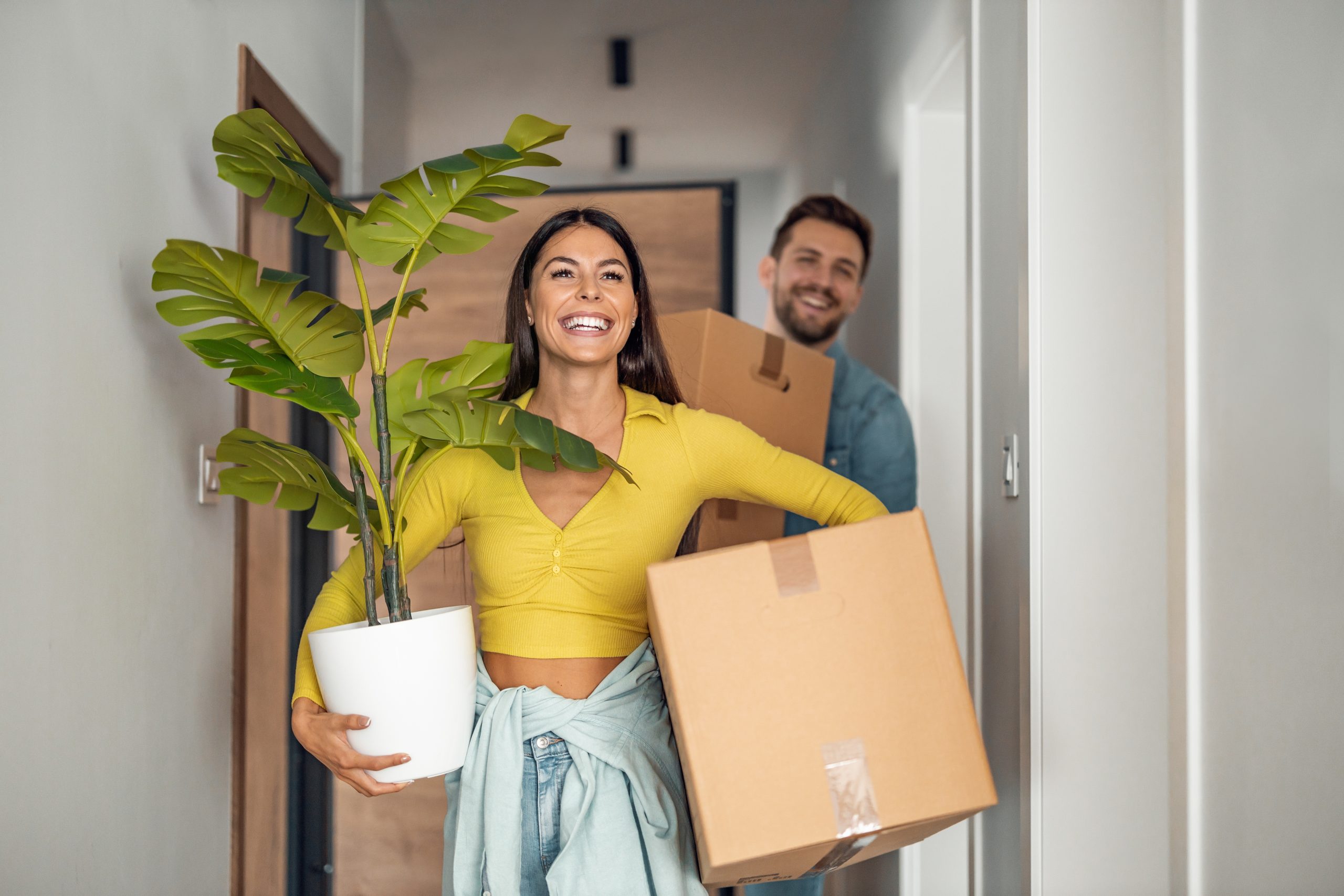 Smiling woman in yellow shirt holding large potted plant in one hand and a box in the other with a man in a blue shirt carrying a box in the background