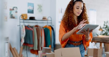 woman standing behind moving boxes looking at ipad. Cover photo for Questions to Ask When Looking for a Commercial Moving Company